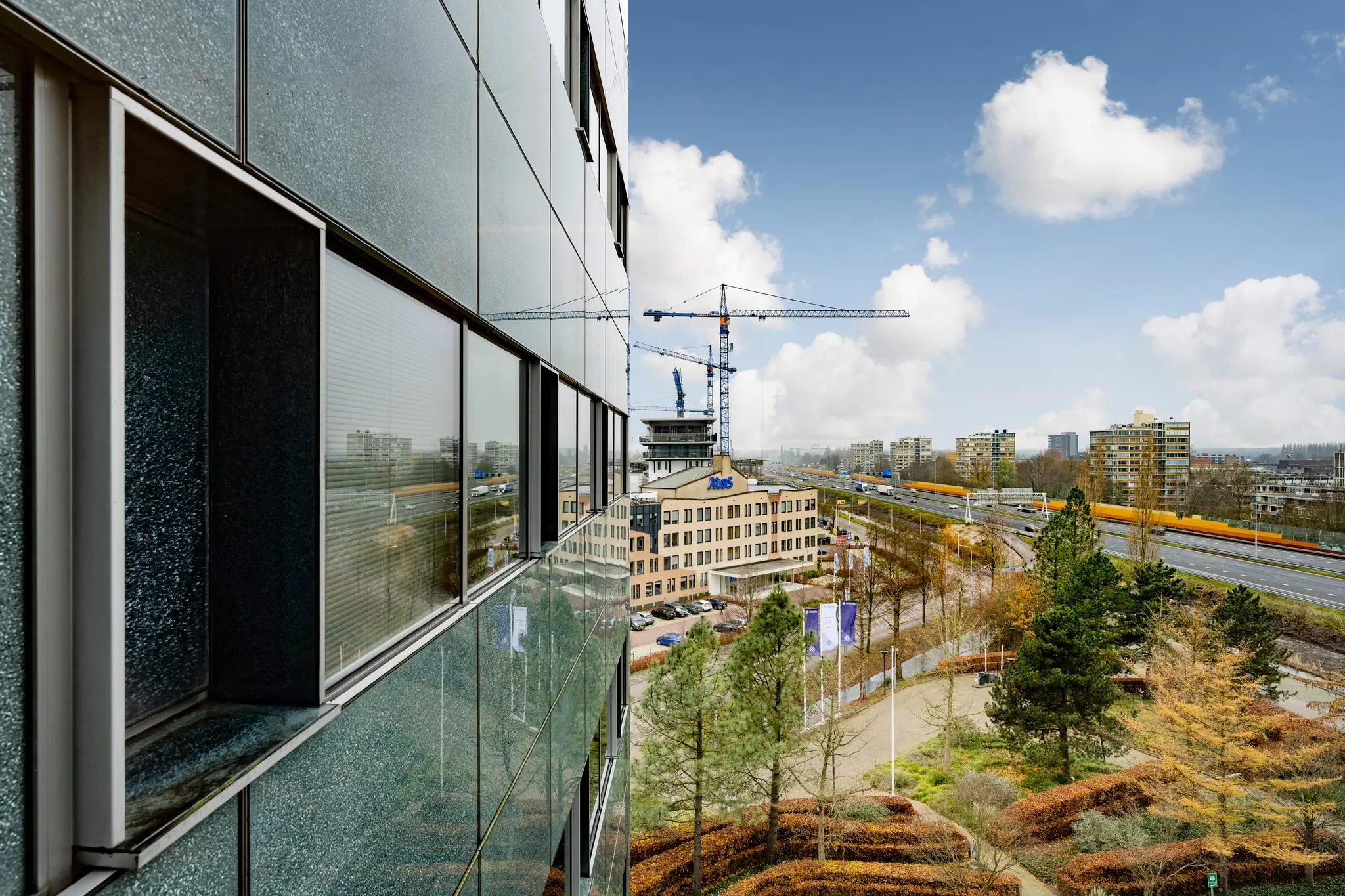 Uitzicht op de Burgemeester Rijnderslaan in Amstelveen met kantoorgebouwen, een bouwkraan en een snelweg met een passerende gele trein.